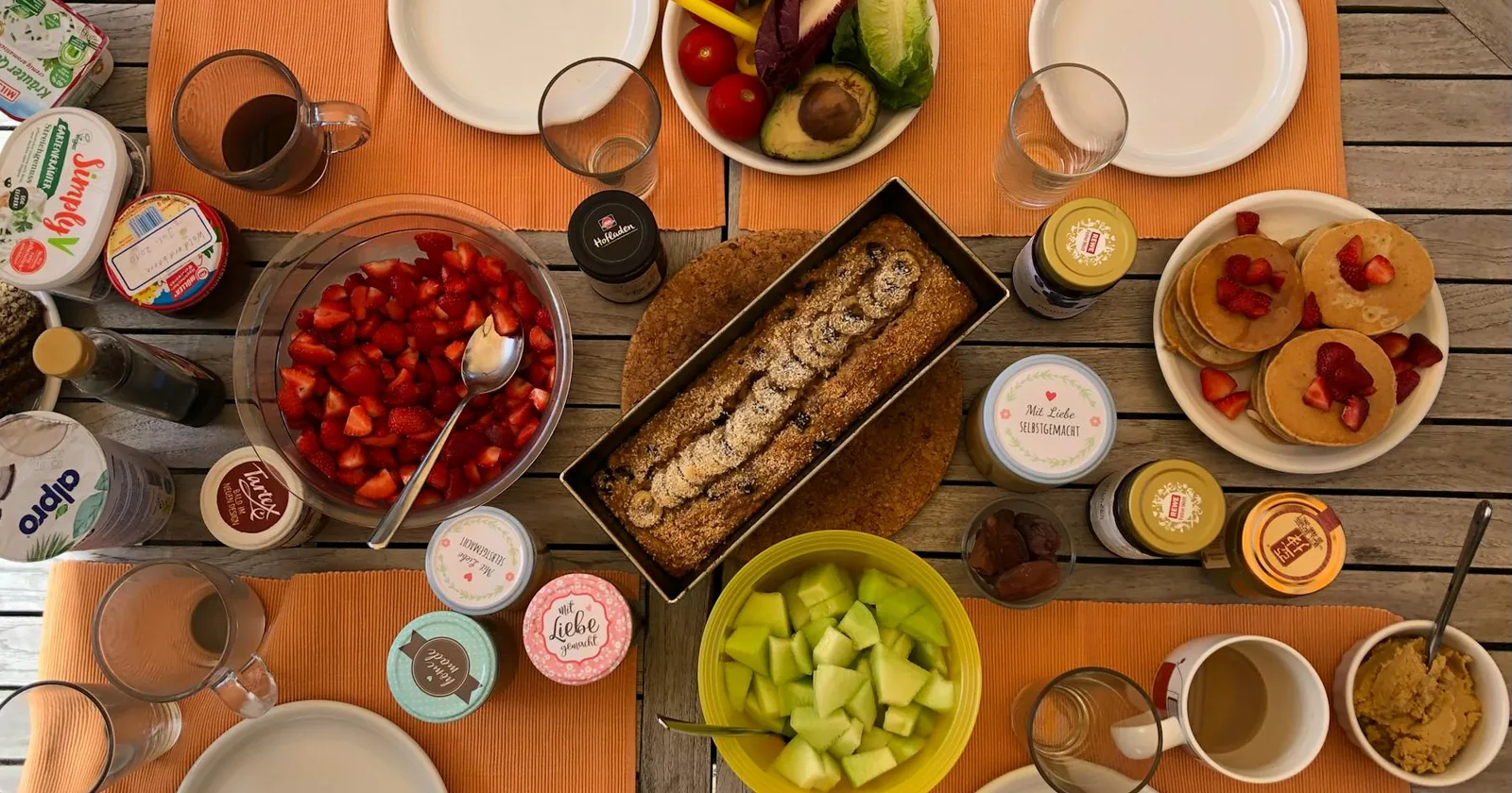 Overhead view of a vegan brunch spread with fruits, pancakes, and bread on a wooden table.