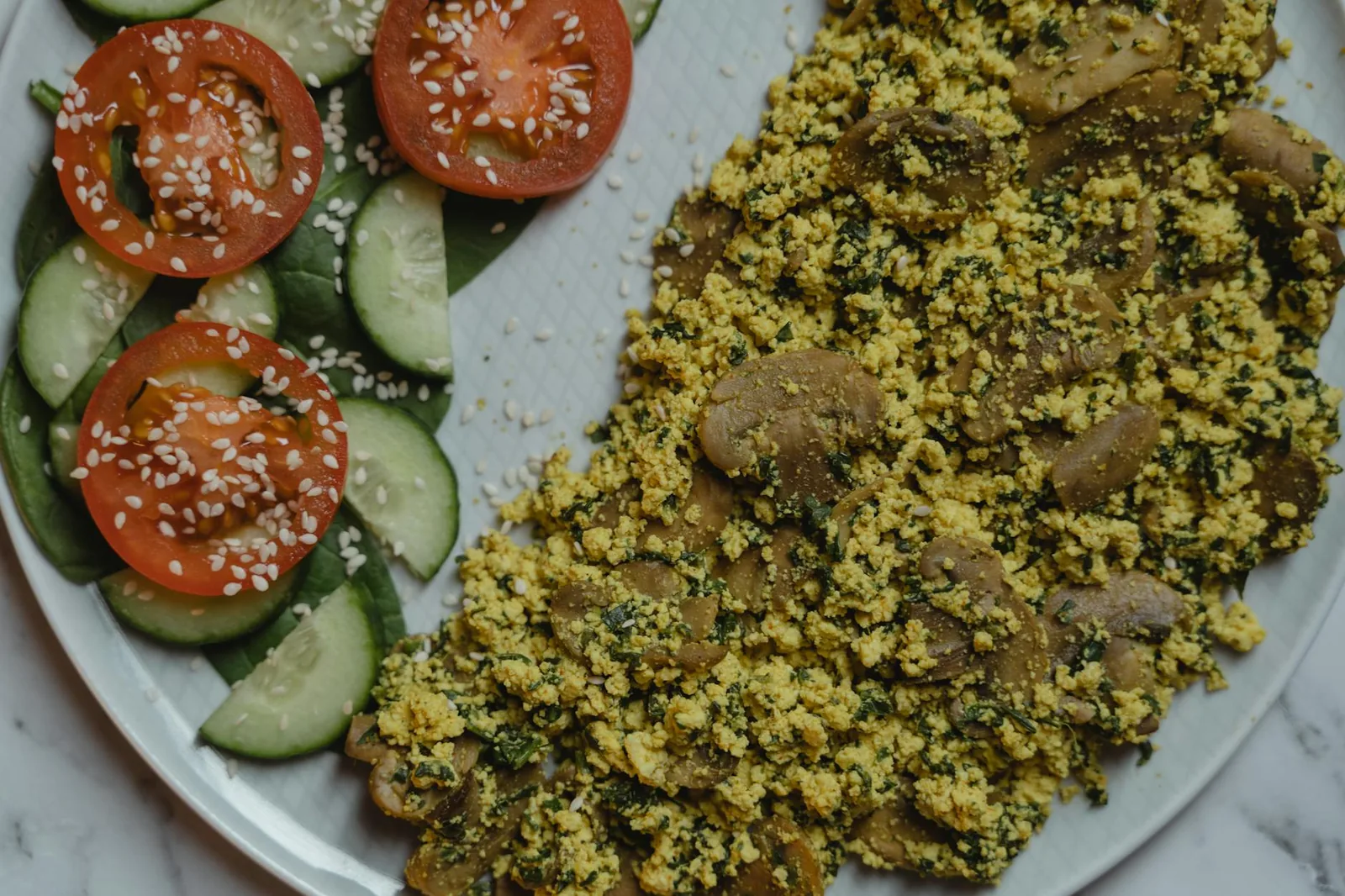 Healthy vegan meal with tofu scramble, cucumbers, and tomatoes on a marble surface.