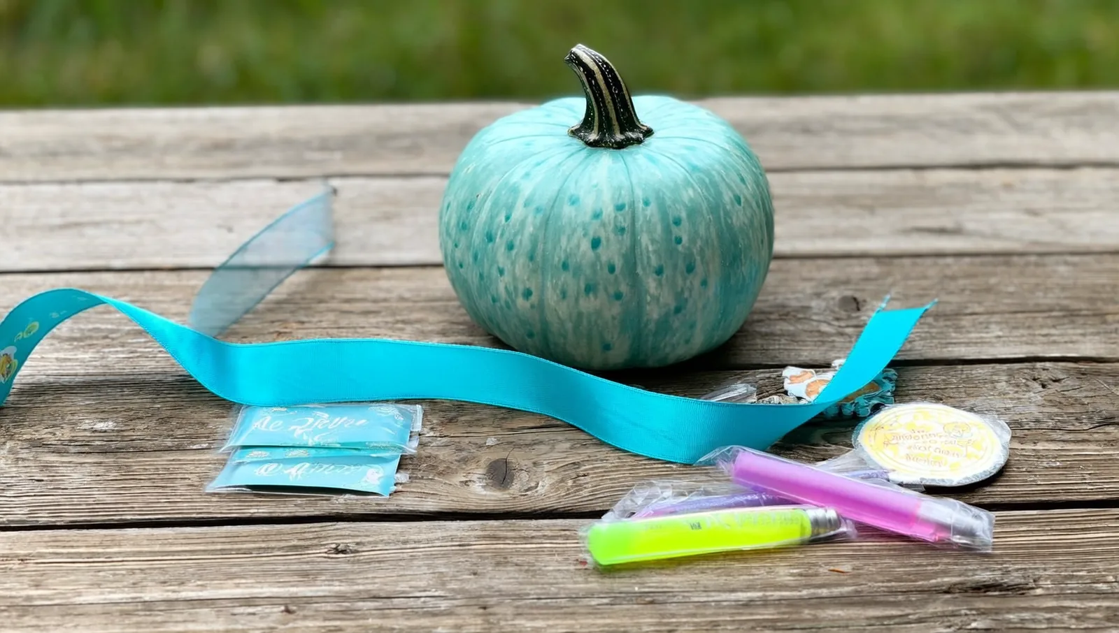 A small teal-painted pumpkin beside a teal awareness ribbon and an assortment of non-food treats on a wooden table.
