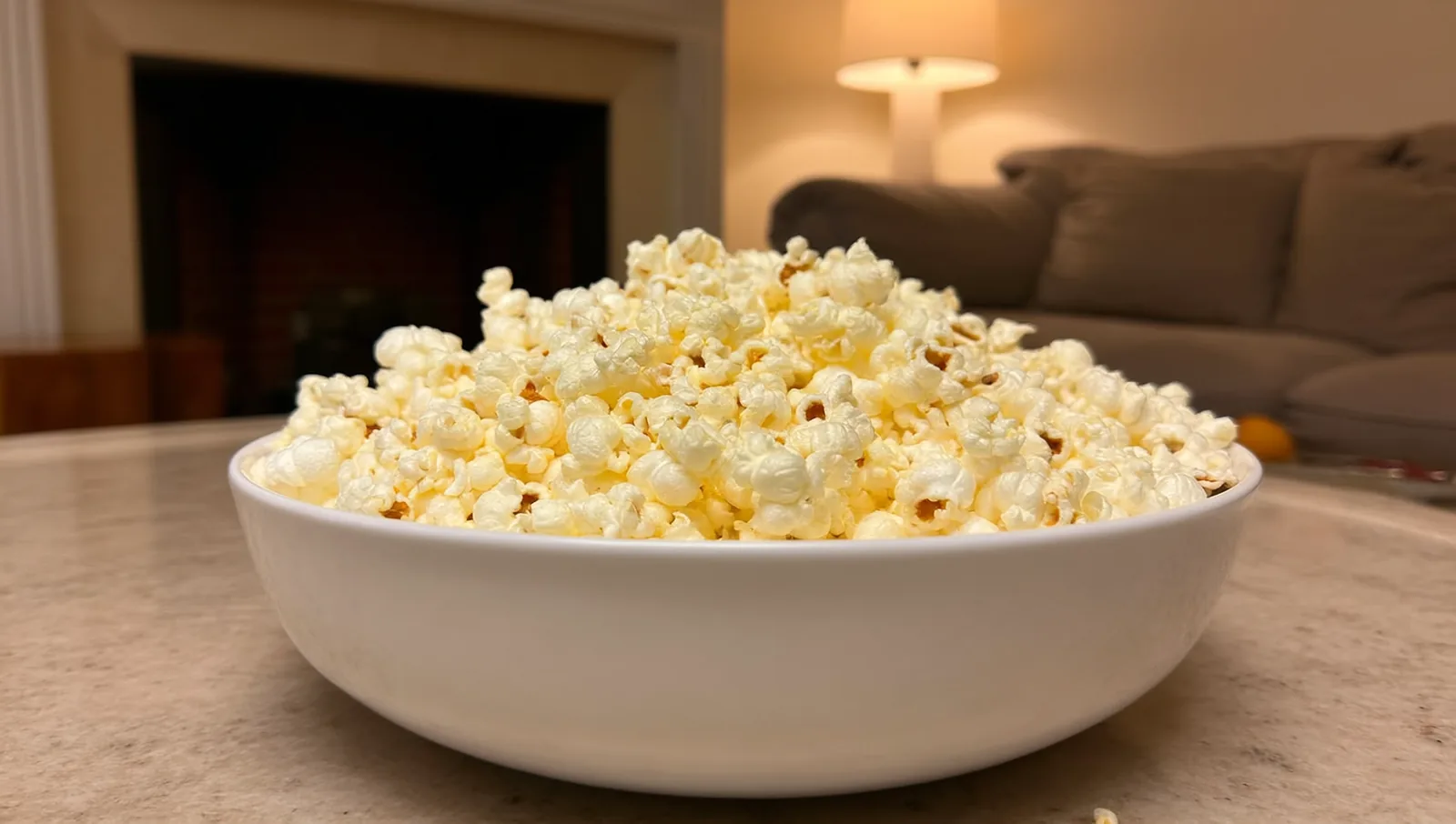 A large white bowl of freshly popped plain popcorn on a table in a cozy living room.