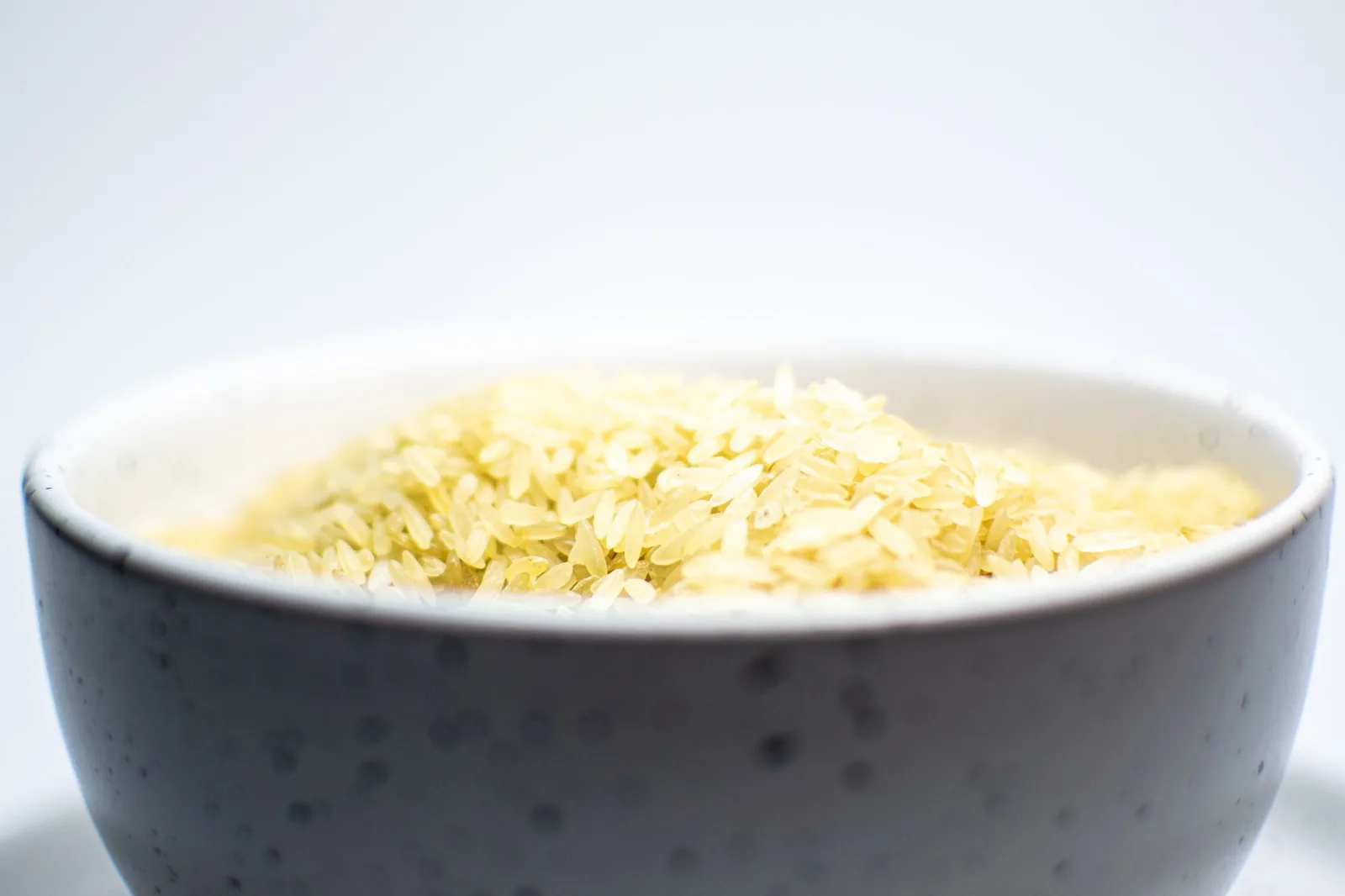 A close-up view of uncooked white rice in a speckled ceramic bowl on a light background.