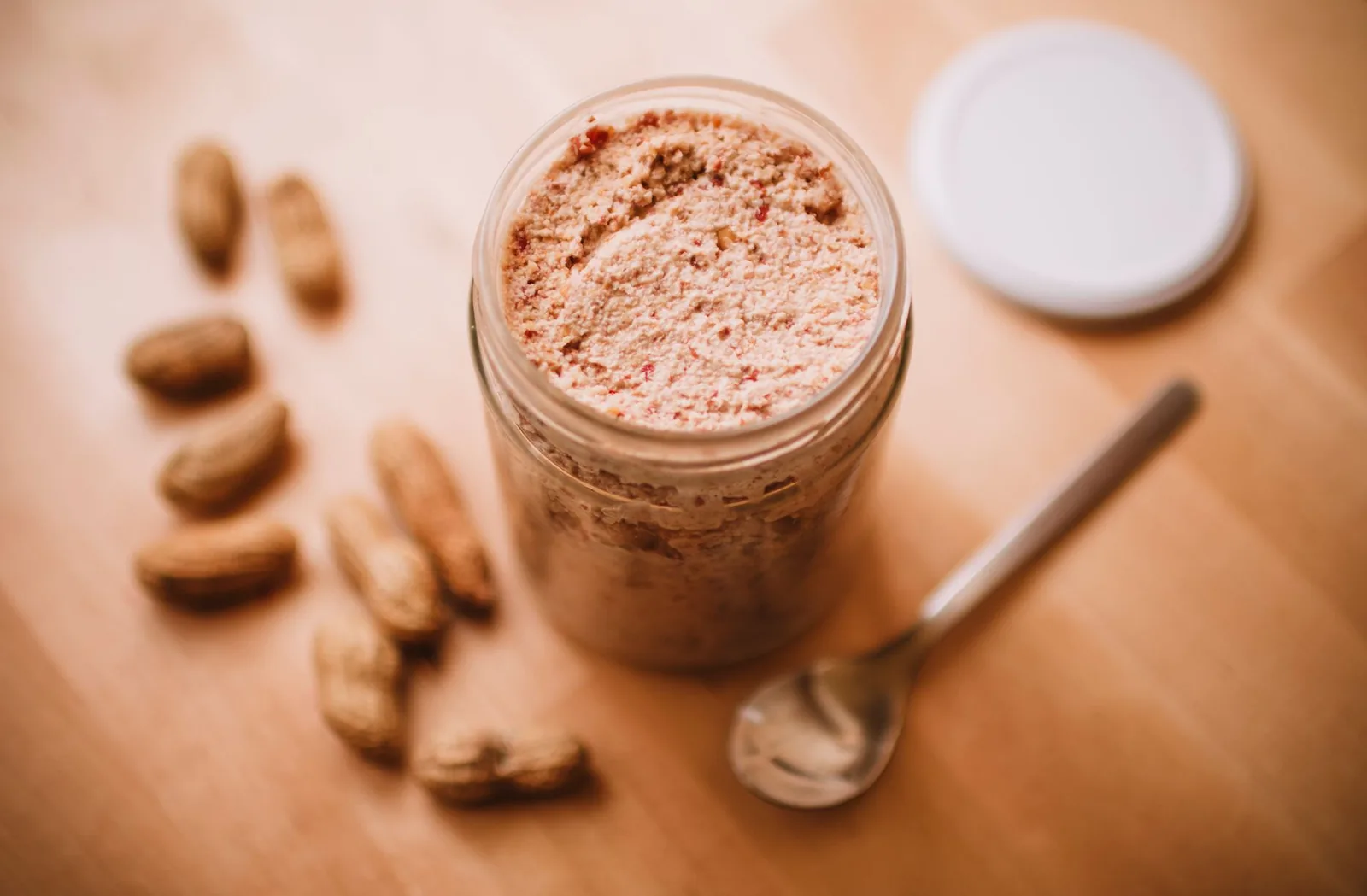 Homemade peanut butter in a glass jar with loose peanuts and a spoon beside it on a table.